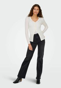 Cardigan bianco aderente a maniche lunghe con scollatura a V e chiusura a bottoni, abbinato a jeans neri a vita alta e stivaletti neri con tacco.