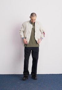 Giacca bomber beige chiaro con polsini e colletto a costine; indossata sopra una maglietta verde oliva. Pantaloni neri e scarpe nere completano l'outfit.