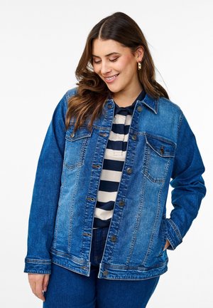 Veste en denim dans différentes nuances de bleu avec une coupe classique, un bouton sur le devant, deux poches poitrine et des manches longues. Portée sur une chemise rayée.