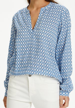 Femme portant un chemisier bleu ample avec un motif losange blanc et des manches longues, associé à un pantalon blanc.