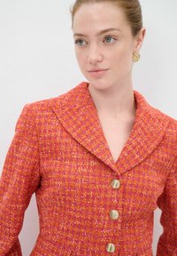 Roter und pinkfarbener strukturierter Blazer mit einem breiten Reverskragen, goldenen Knöpfen und einer taillierten Passform, der ein gewebtes Muster aufweist.