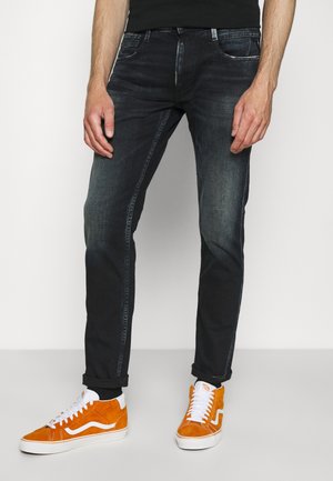 Muž nosící vybledlé černé džíny s vyhrnutými lemy a jasně oranžové semišové tenisky s bílými tkaničkami, stojící proti jednoduchému pozadí.