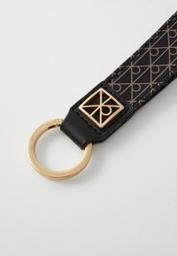 Bracelet porte-clés en cuir noir avec un anneau circulaire doré et un accent de logo carré avec des motifs géométriques sur une surface texturée.