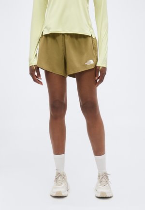 Persona vistiendo pantalones cortos North Face verde oliva, camiseta de manga larga amarillo pálido, calcetines blancos y zapatillas blancas, de pie frente a un fondo liso.