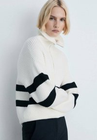 Weißer Strickpullover mit hohem Kragen, ausgestattet mit schwarzen horizontalen Streifen an den Ärmeln. Gerippte Textur, lockere Passform und Reißverschluss am Hals.