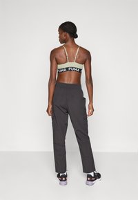 Melnas PUMA zīmola sporta krūšturis ar zaļām siksnām, kombinēts ar tumšiem kargo biksēm. Melni sporta apavi ar rozā akcentiem. Apskats no aizmugures.