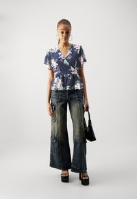 Blomstrete omslagstopp i marineblått med rosa og hvite blomster, kombinert med vide, utvaskede denimjeans. Modellen bærer en liten svart håndveske.