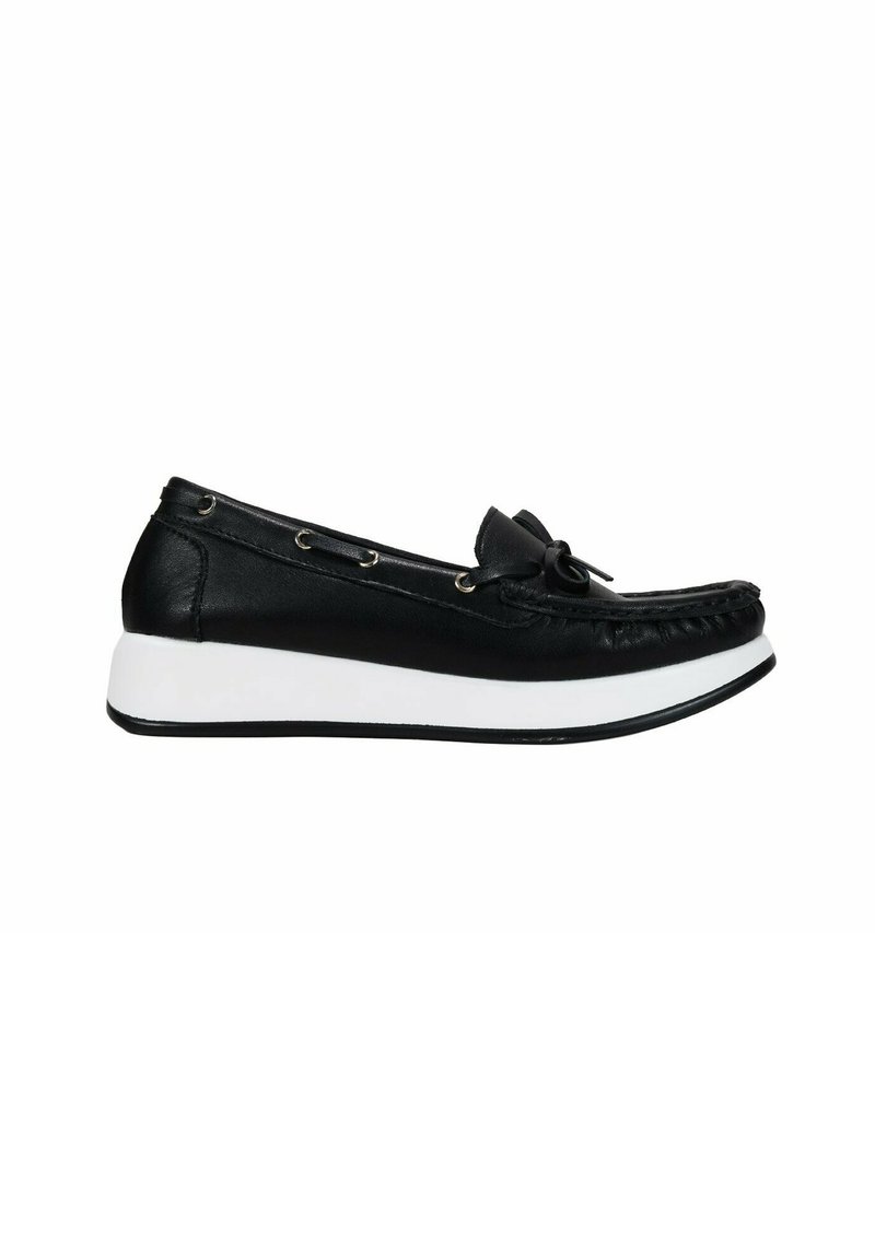 Zapato negro de cuero tipo slip-on con punta redondeada, suela de goma blanca, acento de lazo y ojales decorativos de metal a lo largo del costado.