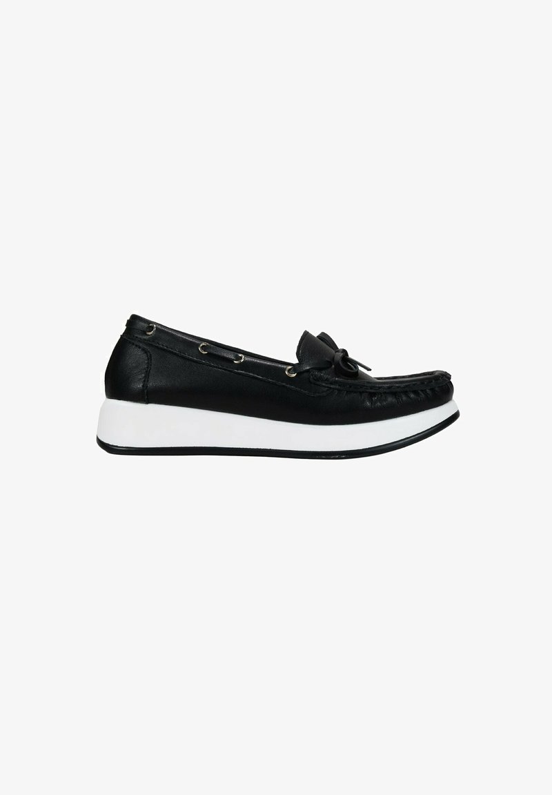 Zapato negro de cuero tipo slip-on con punta redondeada, suela de goma blanca, acento de lazo y ojales decorativos de metal a lo largo del costado.