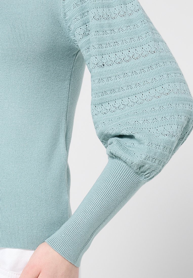 Lichtblauwe pullovertrui met geribde manchetten en pofmouwen, met een gestructureerd patroon van openwerkdetails langs de mouwen.