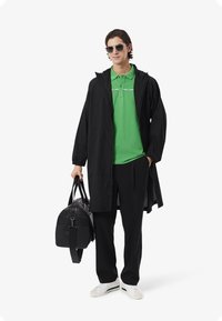 Zwarte lange jas, groen poloshirt met witte strepen, zwarte broek, witte sneakers met groene accenten, draag een zwarte sporttas en een zonnebril.