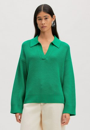 Vrouw met lang donker haar, grote zilveren oorbellen en een groene gebreide trui met V-hals en crèmekleurige broek tegen een effen achtergrond.