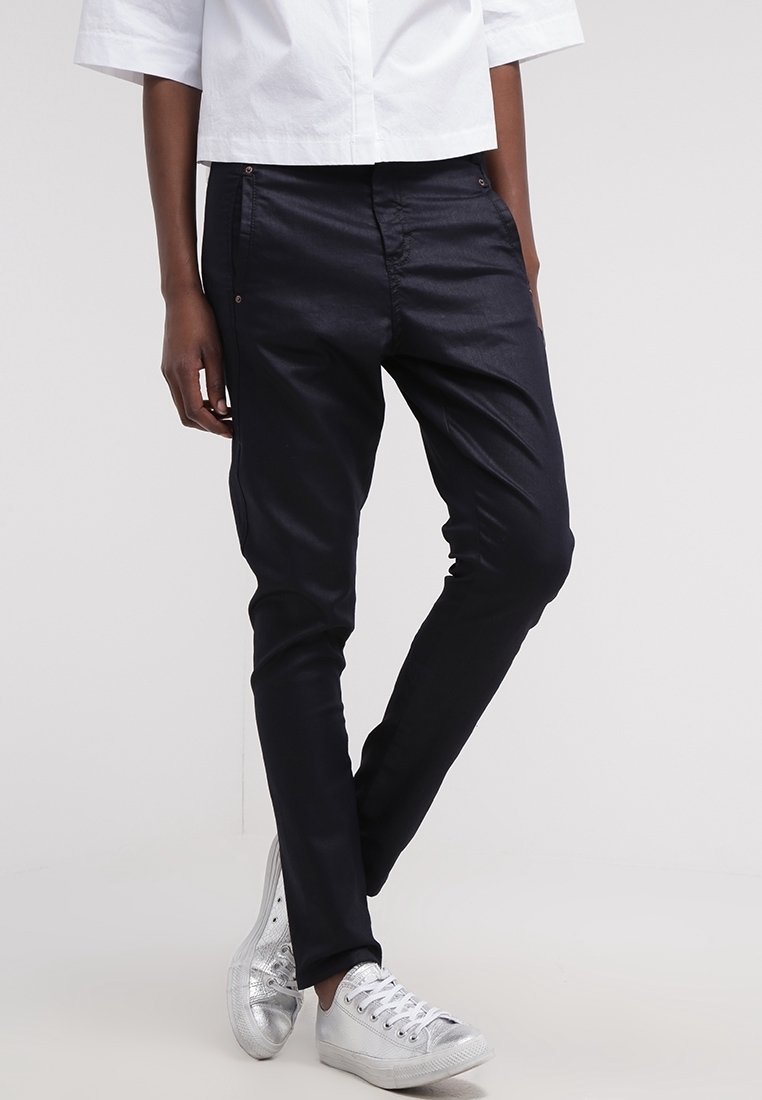 Personne portant un pantalon noir slim fit, une chemise blanche courte et des baskets métalliques argentées, debout avec une jambe croisée devant.