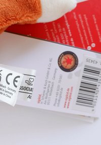 Plush toy with orange fur and white accents, tagged with care instructions and a CE marking. Includes a barcode and logo on the packaging.