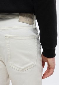 Άτομο που φορά ανοιχτόχρωμο τζιν με μπεζ πατς λογότυπο Calvin Klein στη μέση πίσω και μαύρο μακρυμάνικο τοπ, με το χέρι χαλαρό στο πλάι.