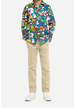 Junge trägt ein buntes langärmliges Hemd mit Super-Mario-Figuren, beigefarbene Hose und weiße Turnschuhe, steht mit den Händen in den Taschen.