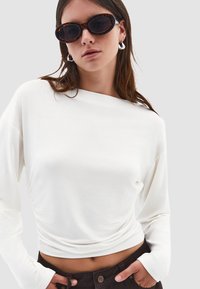 Bílý dlouhý rukáv top s lodičkovým výstřihem, s řasením na boku, doplněný slunečními brýlemi s želvovinovým vzorem a stříbrnými kruhovými náušnicemi.