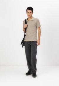 Beige, textúrájú pólóing, galléros nyakkal, sötét szürke, egyenes szárú farmernadrággal és fekete sportcipővel párosítva; a modellen táska van.