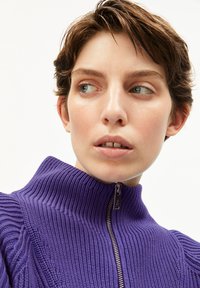 Lila Strickpullover mit hohem Kragen und Reißverschluss. Verfügt über eine gerippte Textur und subtile vertikale Linien. Nahaufnahme des Gesichts des Models.