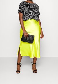 Černý top s krátkým rukávem a bílými abstraktními liniemi, doplněný lesklou neonově žlutou midi sukni s bočním rozparkem; drží černou kabelku.