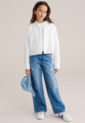 Jauna mergina stovi, dėvi baltą mygtukais užsegamą marškinį, baltą palaidinę, mėlynas plačiomis kojų kelnėmis džinsus, baltus sportinius batelius, šalia laikydama mėlyną kepuraitę.