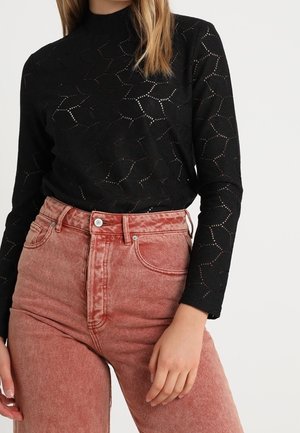 Femme portant un haut noir à manches longues à motifs, rentré dans un jean rouge délavé taille haute, sur un fond uni.