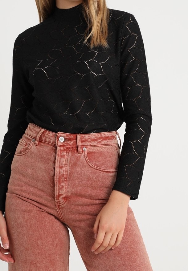 Femme portant un haut noir à manches longues à motifs, rentré dans un jean rouge délavé taille haute, sur un fond uni.