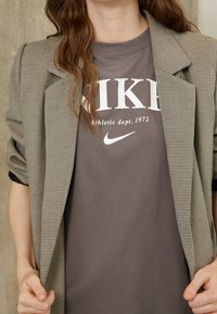 Blazer beige a motivo houndstooth sopra una t-shirt grigia Nike con lettere bianche. Il blazer ha spalle strutturate e maniche arrotolate.