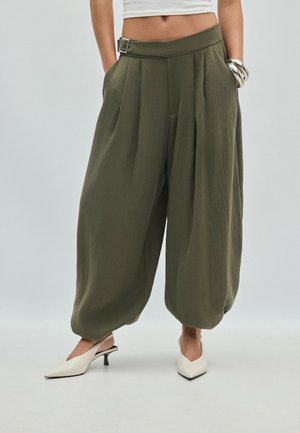 Donna con pantaloni larghi verde oliva con tasche laterali, top bianco corto, braccialetti d'argento e tacchi slingback bianchi con tacco basso.