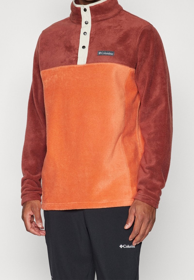 Sudadera de forro polar con cuello alto, que presenta un diseño en dos tonos en óxido y naranja, botones a presión y un logo de Columbia en el pecho.