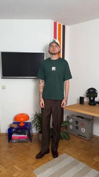 Άνδρας με τατουάζ, φορώντας σκούρο πράσινο πουκάμισο και καπέλο μπέιζμπολ, στέκεται σε δωμάτιο με τηλεόραση τοποθετημένη στον τοίχο, πολύχρωμες ρίγες, φυτά και γραφείο.