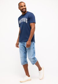 Mörkblå bomullströja med "MUSTANG" tryckt i vitt, kombinerat med ljusblå knälånga jeansshorts och vita sneakers.