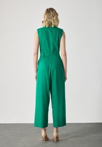 Green sleeveless jumpsuit with a belt, wide-leg design, and textured fabric, paired with beige high-heeled shoes.