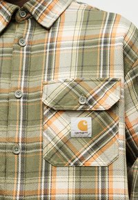 Πράσινο, πορτοκαλί και κρεμ πουκάμισο καρό με κουμπιά, με μπροστινή τσέπη που διαθέτει καπάκι με κουμπί και ένα λογότυπο Carhartt.