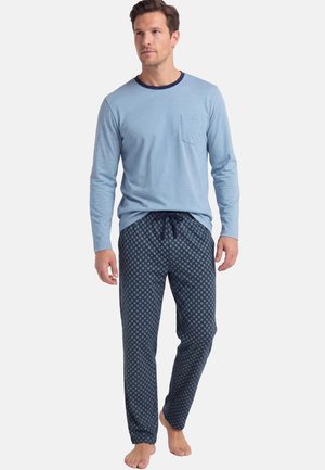 Uomo in piedi a piedi nudi che indossa una maglia a maniche lunghe azzurro chiaro con taschino sul petto e pantaloni del pigiama scuri con motivo e coulisse.