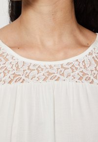 Lättviktstopp i vitt med en blommig spetsdetalj på axlarna, rundad halsringning och veckad textur under bröstet.