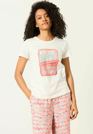 Kobieta z kręconymi włosami, ubrana w białą koszulkę z grafiką śpiących sardynek oraz różowe spodnie w wzory z niebieskimi rybami i czerwonymi falowanymi liniami.