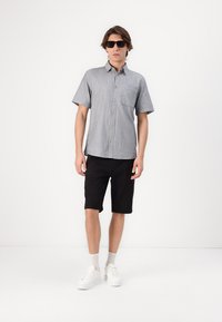 Chemise à manches courtes pour homme en gris avec une poche, associée à un short noir et des baskets blanches, se tenant sur un fond blanc.