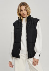 Juodas paminkštintas liemenė su auk�štu apykakle, be rankovių dizainu ir priekiniu užtrauktuku. Dėvima virš baltos banguotos megztinės. Lygus paviršius.