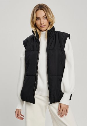 Gilet rembourré noir avec un col montant, sans manches, et fermeture éclair à l'avant. Porté par-dessus un pull à côtes blanc. Texture lisse.