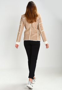 Woman walking away wearing a beige jacket with white trim, black pants, and silver sneakers against a plain light background.