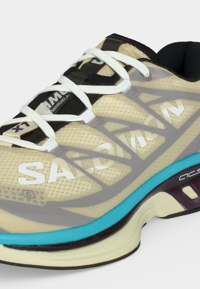 Μπεζ και μαύρο αθλητικό παπούτσι με λευκά κορδόνια, μπλε λεπτομέρεια στη μεσαία σόλα και σήμανση "SACAI" στο πλεκτό επάνω μέρος και στην μαύρη ετικέτα της γλώσσας.