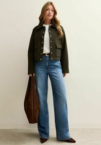 Groene cropped jas met gouden knopen, witte blouse en uitlopende blauwe spijkerbroek. Bruine tas in de hand, puntige schoenen maken de look af.