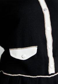 Närbild av ett svart tygplagg med vit knäppkant, en vit knappficka med lock och dekorativa pärlkanter.