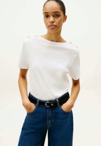 Hvit kortermet skjorte med båthals og perlepynt på skuldrene, kombinert med høyt belte blå jeans og et svart belte.