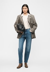 Γυναίκα που φοράει ένα καρό oversized σακάκι, μπεζ μπλούζα, μπλε τζιν, καφέ μπότες, κρατώντας μια μαύρη δερμάτινη τσάντα φάκελο σε λευκό φόντο.