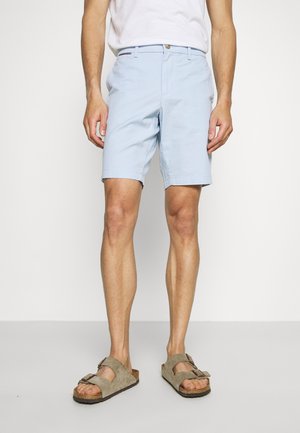 Mann trägt hellblaue Shorts, weißes T-Shirt und beige Wildledersandalen und steht vor einem schlichten weißen Hintergrund.