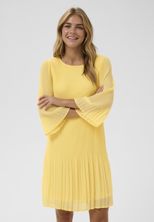Młoda kobieta uśmiechająca się z założonymi na siebie ramionami, ubrana w plisowaną żółtą sukienkę z rozkloszowanymi, półprzezroczystymi rękawami na jednolitym tle.