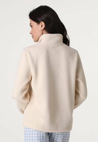 Giacca in pile beige con collo alto, caratterizzata da una superficie morbida e strutturata e da una vestibilità comoda. Indossata con pantaloni a quadretti blu e bianchi.