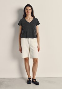 Ženska stoji ob prazni steni, oblečena v črno majico z V-izrezom, bele kratke hlače do kolen in črne sandale s pokritimi prsti.